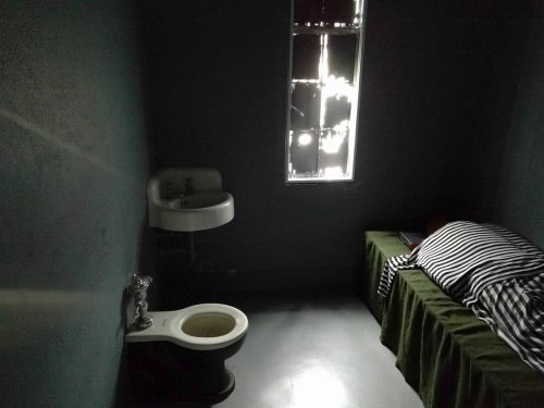 A dimly lit prison cell with a bed, sink, and toilet, featuring a barred window letting in faint light.