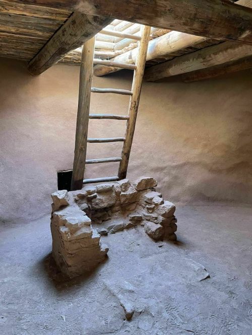 Interior of a rustic room with a stone hearth and a wooden ladder leading to a light source above.