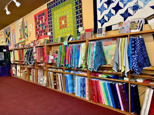 A colorful display of quilts and fabric rolls on shelves in a craft store, with a vibrant carpeted floor.