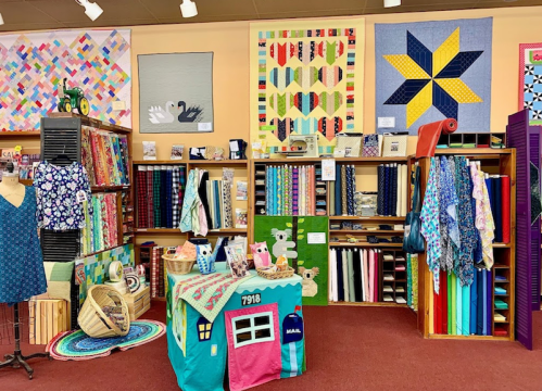 A colorful fabric store display featuring quilts, fabrics, and sewing supplies arranged on shelves and tables.