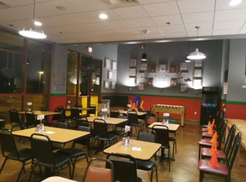 A brightly lit restaurant interior with empty tables, chairs, and wall decor, including framed pictures and an arcade game.
