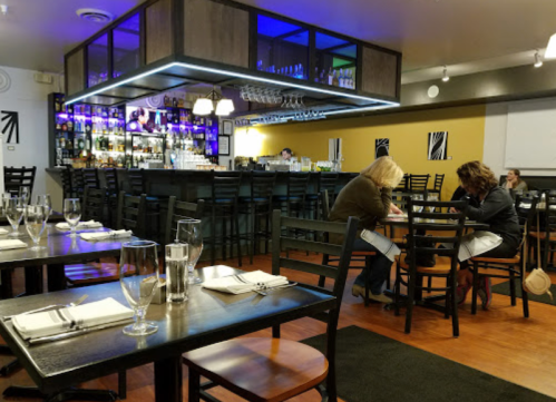 A modern restaurant interior with a bar, empty tables, and two people seated at a table, engaged in conversation.
