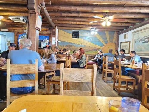 A rustic restaurant interior with wooden tables and chairs, featuring patrons dining and a mural of a sunset on the wall.