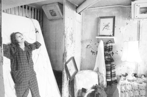 A person in pajamas stands with arms outstretched in a dimly lit, worn room with peeling walls and scattered furniture.