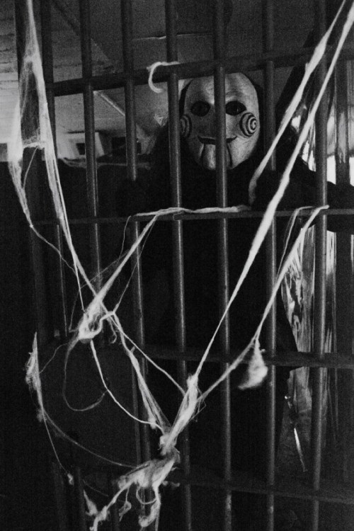 A figure in a creepy mask stands behind prison bars, surrounded by cobwebs in a dimly lit setting.
