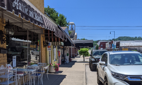 A sunny street scene featuring shops, outdoor seating, and parked cars in a small town.