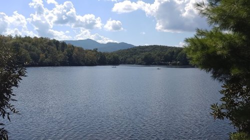 A serene lake surrounded by trees and mountains, with a few kayakers on the water under a partly cloudy sky.