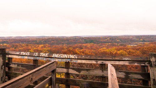 View from a wooden lookout tower overlooking a vibrant autumn landscape with colorful trees and a cloudy sky.