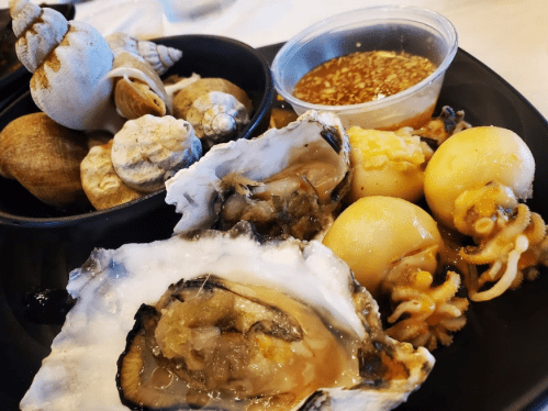 A black plate with oysters, small octopuses, and various shellfish, accompanied by a dipping sauce.