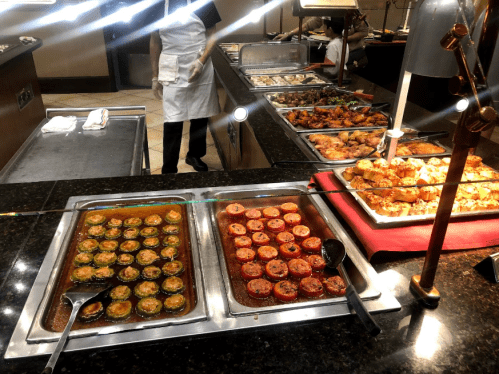 A buffet display featuring various dishes, including baked goods and colorful entrees, with a server in the background.