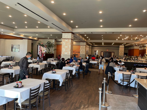 A spacious restaurant with white tablecloths, diners seated at tables, and a buffet area in the background.