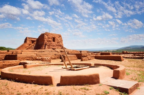 Ancient adobe ruins with a circular structure in the foreground, set against a blue sky with scattered clouds.