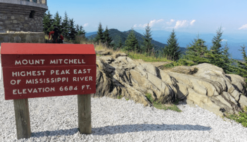 Sign marking Mount Mitchell, the highest peak east of the Mississippi River, with scenic mountain views in the background.