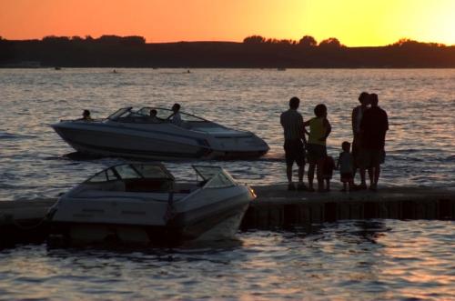 Silhouetted figures stand by the water at sunset, with boats nearby and a calm lake reflecting the orange sky.