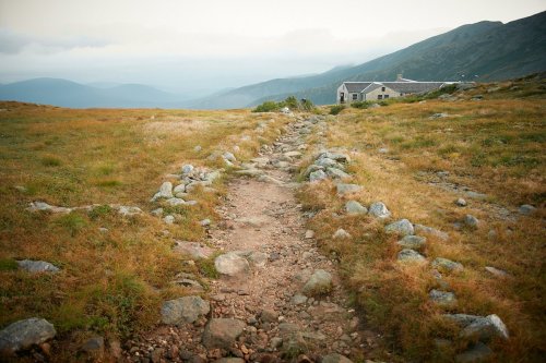 A rocky path leads through grassy terrain towards a distant building, surrounded by mountains under a cloudy sky.