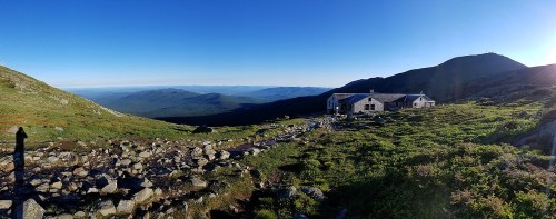 A panoramic view of a mountain landscape with a building, rocky terrain, and distant hills under a clear blue sky.