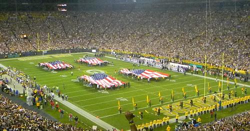 A packed stadium with large American flags on the field during a football game, surrounded by cheering fans.