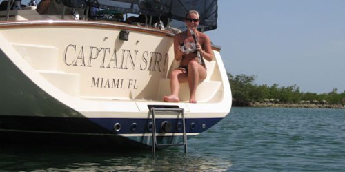 A woman sits on the back of a boat named "Captain Sir" in Miami, holding a small fish, with water in the background.