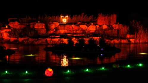 A dark scene featuring a glowing rock formation and colorful lights reflecting on water, creating a festive atmosphere.