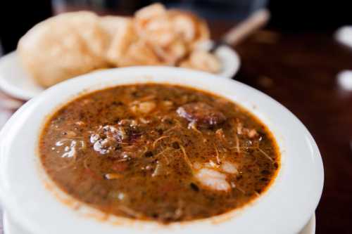 A bowl of rich, dark soup with shrimp and meat, served alongside crispy bread on a wooden table.