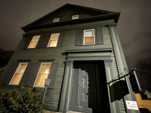 A tall green house at night, featuring dark shutters and illuminated windows, with a sign near the entrance.