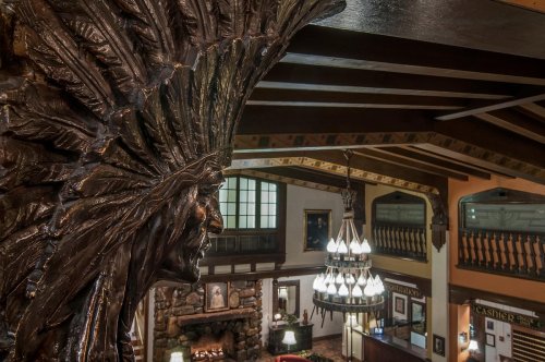 A carved Native American figure overlooks a rustic lodge interior with wooden beams and a chandelier.