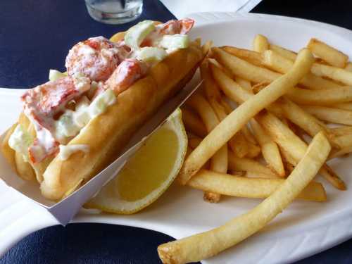 A lobster roll topped with mayonnaise and celery, served with a side of golden French fries and a lemon wedge.