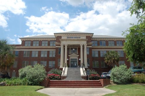 Historic brick building with columns, stairs, and landscaped greenery under a partly cloudy sky.