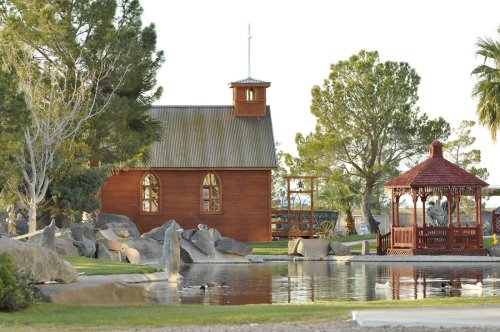 A wooden chapel with a steeple beside a gazebo, surrounded by trees and a pond with rocks in a serene outdoor setting.