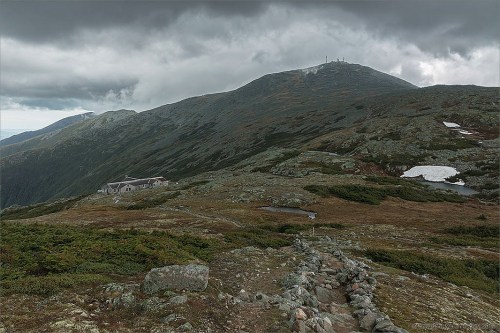 A rocky landscape with a mountain in the background, under a cloudy sky, featuring a small building on the slope.