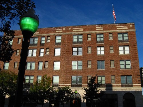 A brick building with multiple windows and an American flag on top, alongside a green streetlamp and trees.