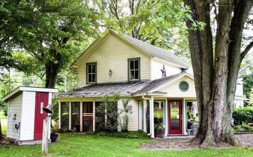A charming two-story house surrounded by trees, featuring red doors and a small shed nearby.