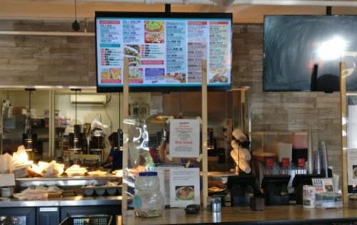 Menu board displayed on a screen in a restaurant, with various food options listed. Kitchen area visible in the background.