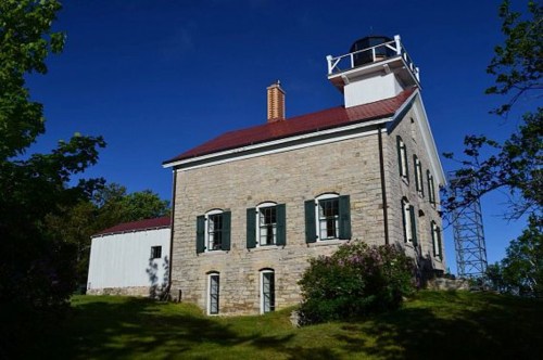 A historic stone lighthouse with a red roof, surrounded by greenery and a clear blue sky.
