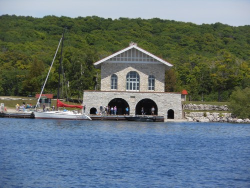 A stone building by the water with people gathered outside, surrounded by green hills and a sailboat nearby.