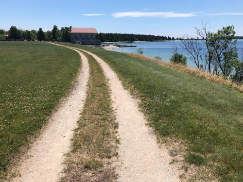 A dirt path leads through green grass to a calm waterfront, with a building in the background under a clear blue sky.