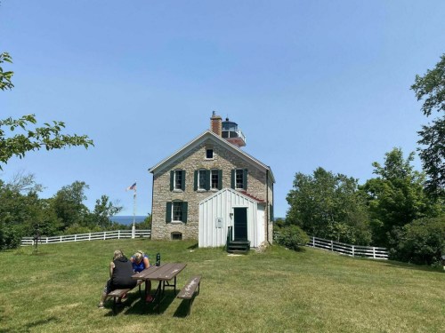 A stone lighthouse with a white roof, surrounded by greenery, with two people sitting at a picnic table in front.