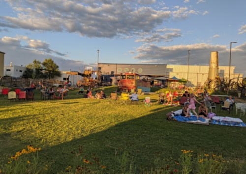 A lively outdoor scene with people sitting on chairs and blankets in a grassy area, surrounded by buildings and trees.
