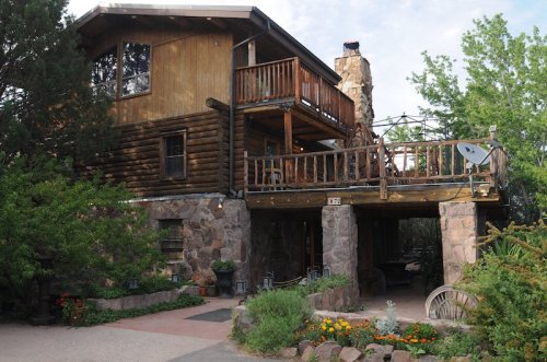 A rustic log cabin with a stone foundation, featuring a balcony and surrounded by greenery and colorful flowers.