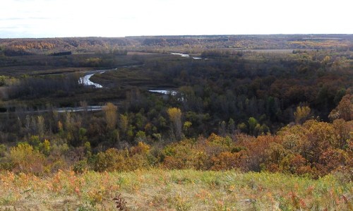 A scenic view of a winding river surrounded by autumn foliage and rolling hills under a partly cloudy sky.