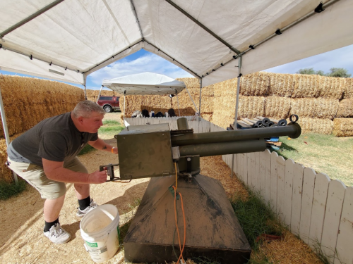 A man aims a cannon-like device under a tent, surrounded by hay bales and equipment.