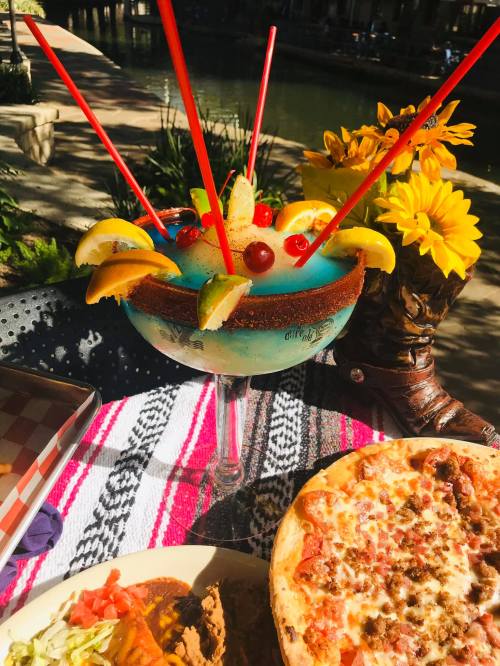 A colorful drink with fruit garnishes and straws, alongside a pizza and a plate of food on a vibrant table setting.