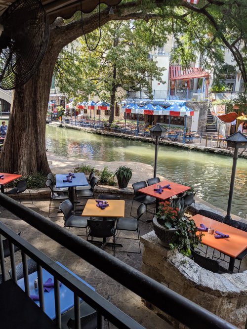 Outdoor dining area by a canal, featuring colorful tables and chairs, surrounded by trees and buildings.