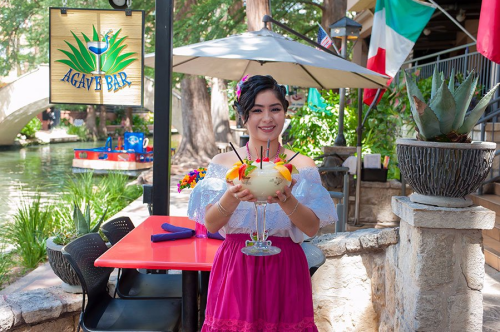 A woman in traditional attire holds a colorful drink at Agave Bar, with flags and a scenic river in the background.