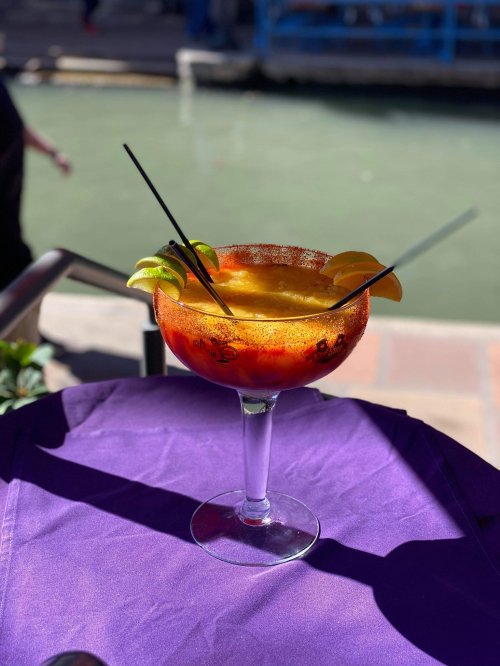 A vibrant cocktail in a large glass, garnished with lime and orange slices, sits on a purple tablecloth.