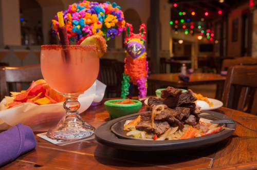 A vibrant table setting featuring a colorful piñata, a cocktail, and a sizzling platter of steak with vegetables.