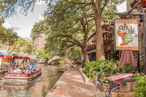 A scenic view of a riverwalk with boats, trees, and a colorful café sign in a vibrant outdoor setting.
