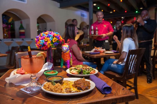 A vibrant restaurant scene with colorful decor, featuring a table of Mexican dishes and drinks, and musicians performing nearby.