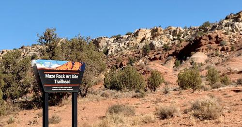 Sign for Maze Rock Art Site Trailhead in a desert landscape with rocky hills and sparse vegetation. Clear blue sky above.
