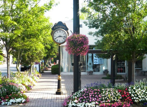 A charming street scene with a clock, colorful flower beds, and trees lining a walkway in a quaint town.